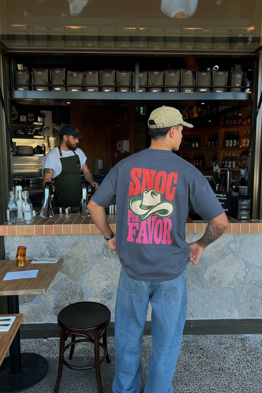 Camiseta SNOC Por Favor vista por la espalda con el sombrero vaquero y el lettering completo, en la barra de un café