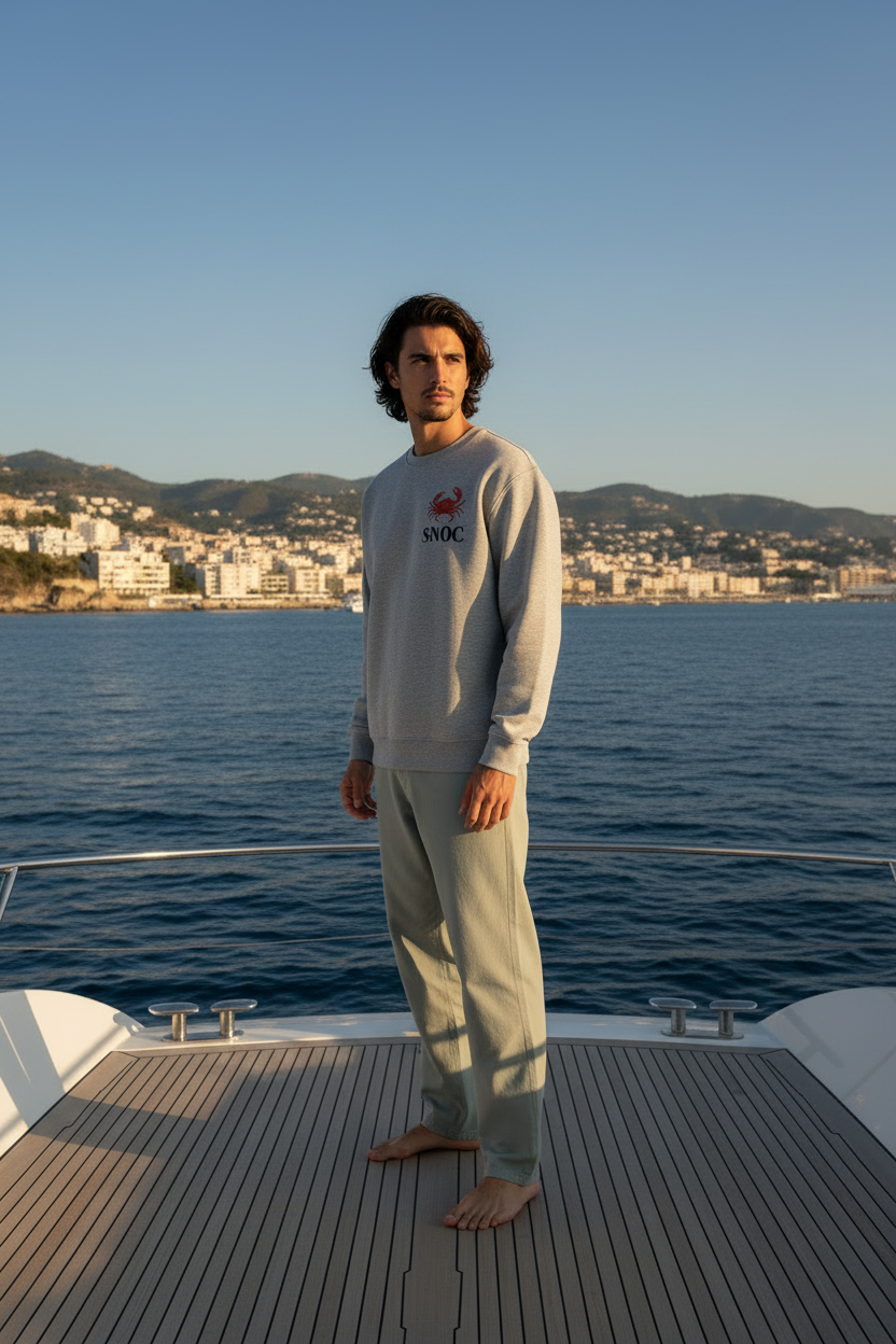 Chico con la sudadera Crab and Sea de SNOC en gris jaspeado, vista frontal con el sello del cangrejo en el pecho, junto a un yate