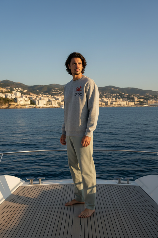Chico con la sudadera Crab and Sea de SNOC en gris jaspeado, vista frontal con el sello del cangrejo en el pecho, junto a un yate